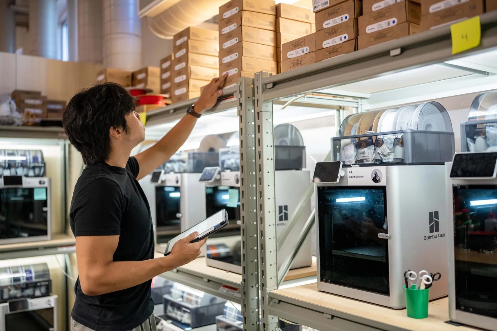 Jr Perez checking inventory on a shelf of 3D printers, tablet in hand